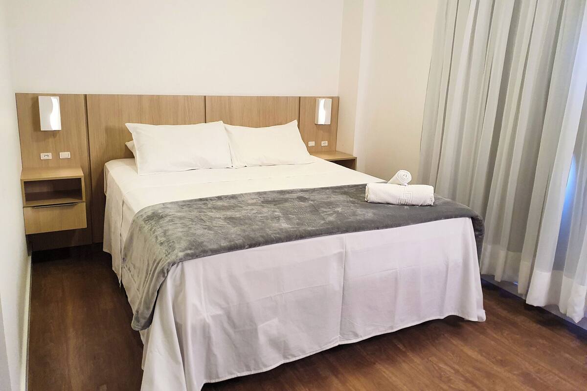 Una cama bien hecha con sábanas blancas, un edredón gris y una toalla enrollada al lado. Hay dos mesitas de noche con lámparas, y una ventana con cortinas.