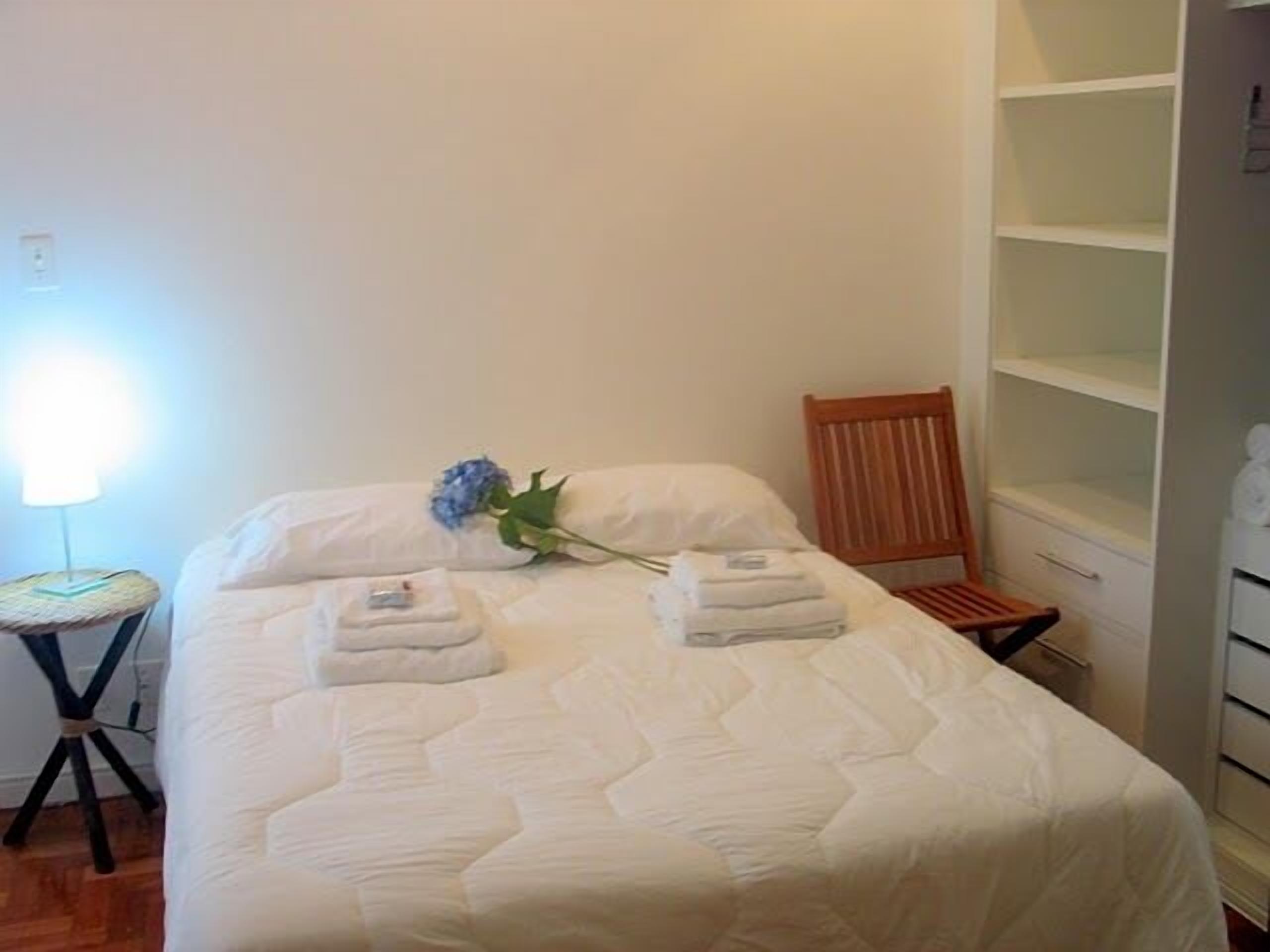 A neatly made bed with a blue flower and folded towels, a wooden chair, and a white wardrobe.