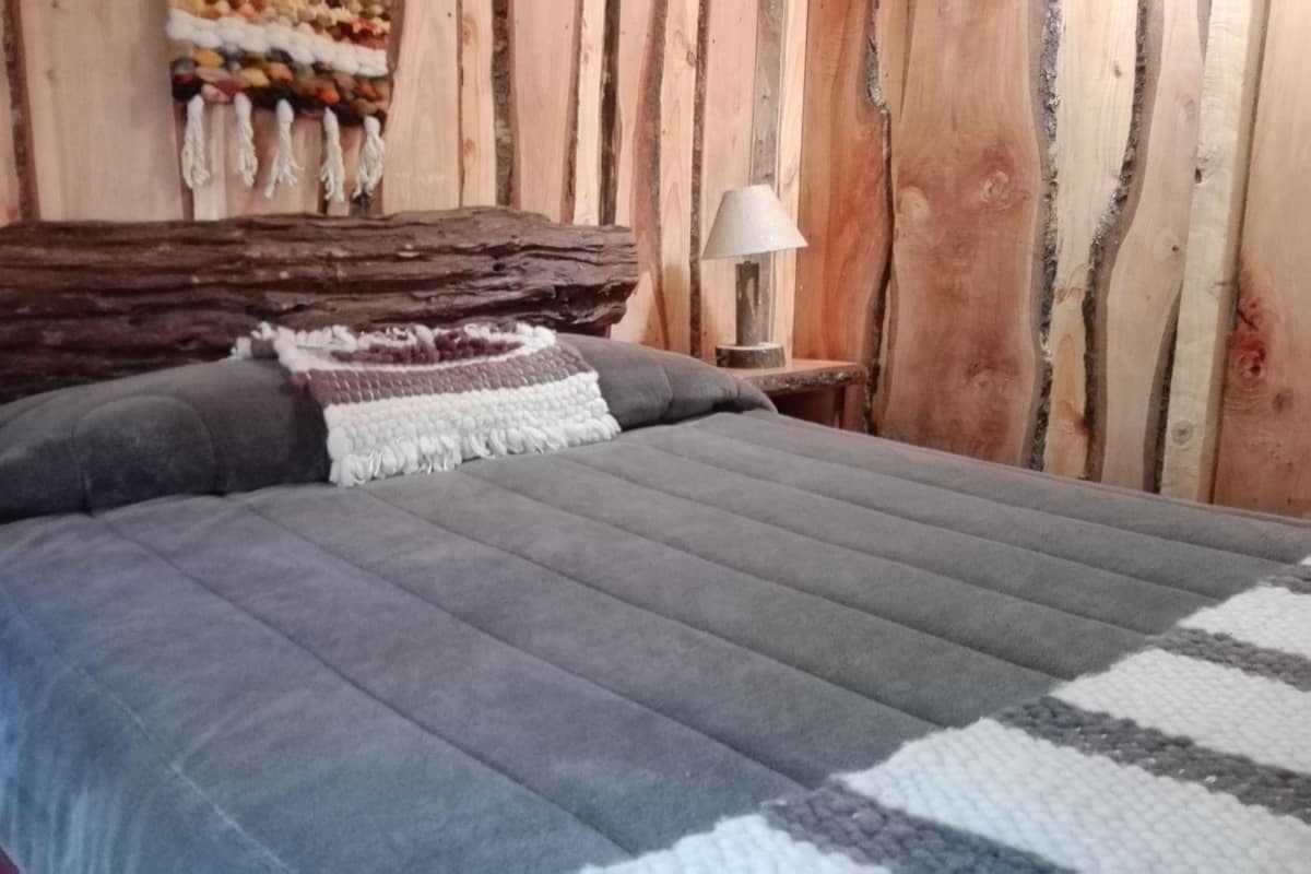 A bed with a grey blanket and pillows, a wooden headboard, a lamp on a nightstand, and a wall with vertical wooden planks.