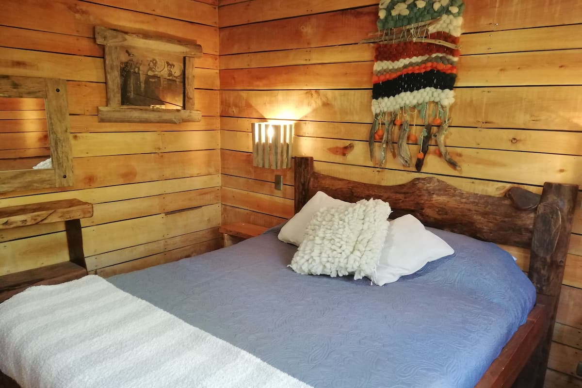 A wooden cabin room with a bed, a nightstand, a wall-mounted lamp, and a decorative wall hanging.