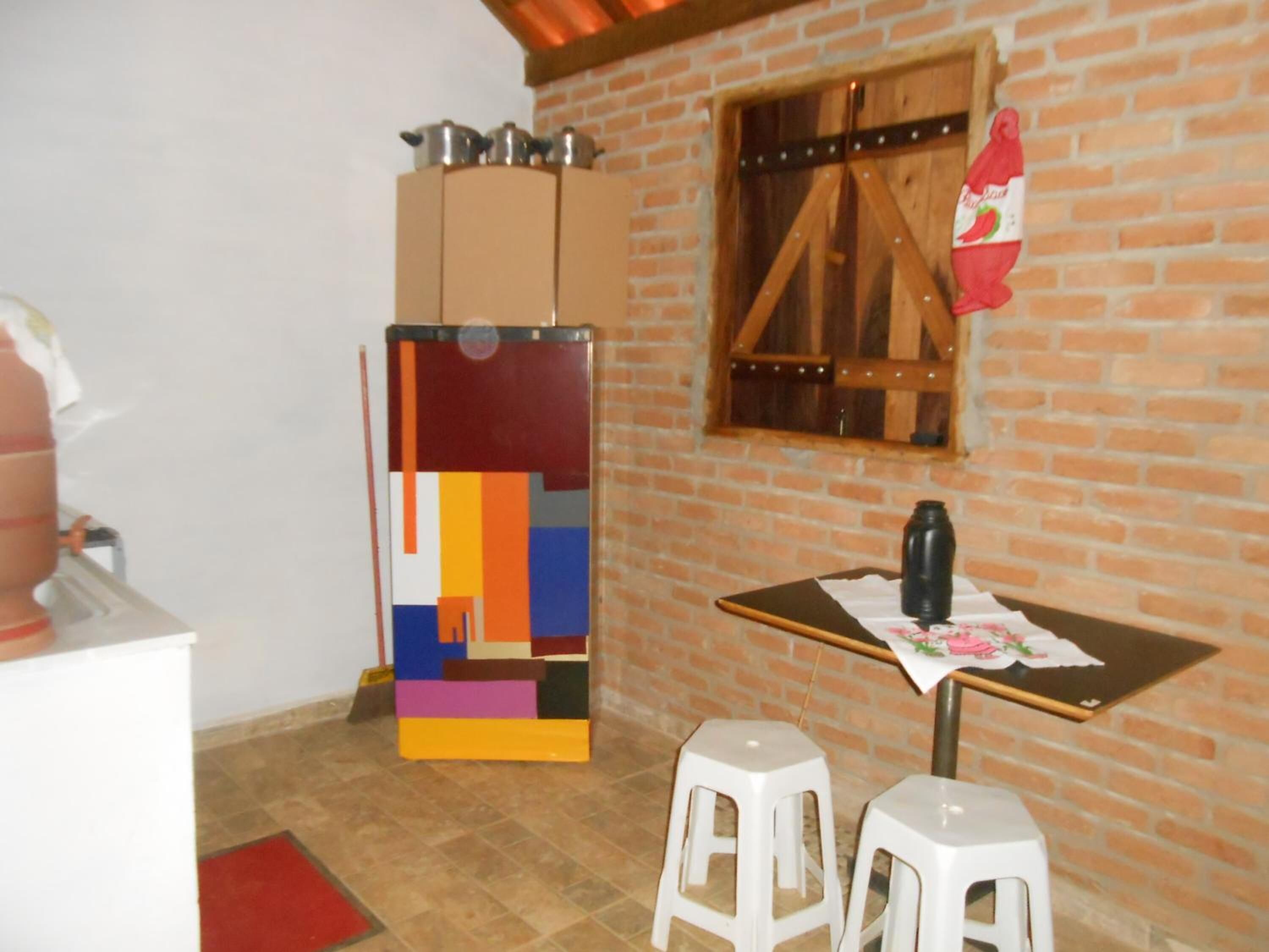 Una habitación pequeña con pared de ladrillo, ventana de madera, una mesa con una botella y dos taburetes.