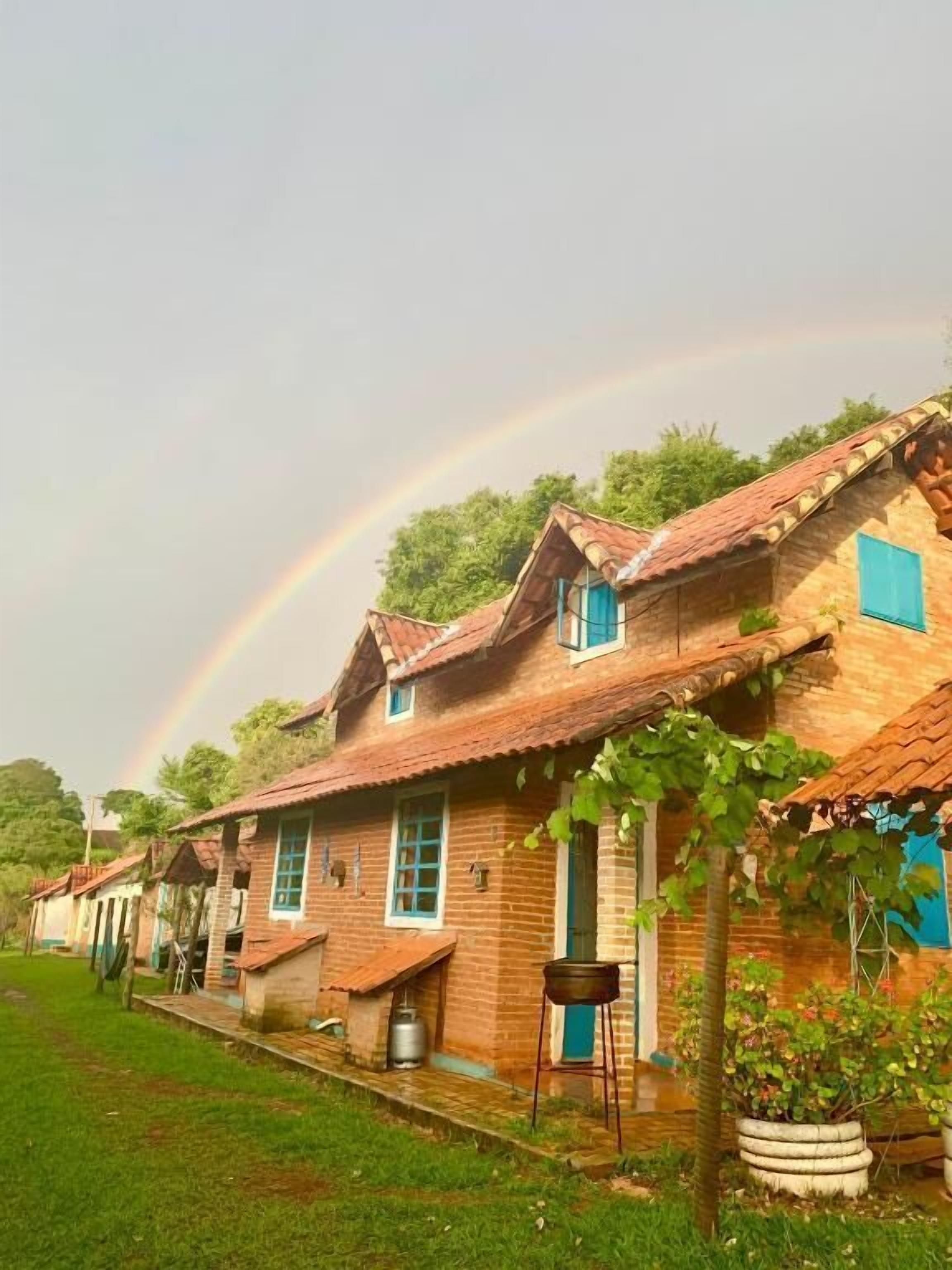 A row of traditional brick houses with pitched roofs and blue shutters, a grassy lawn, and a rainbow in the sky.