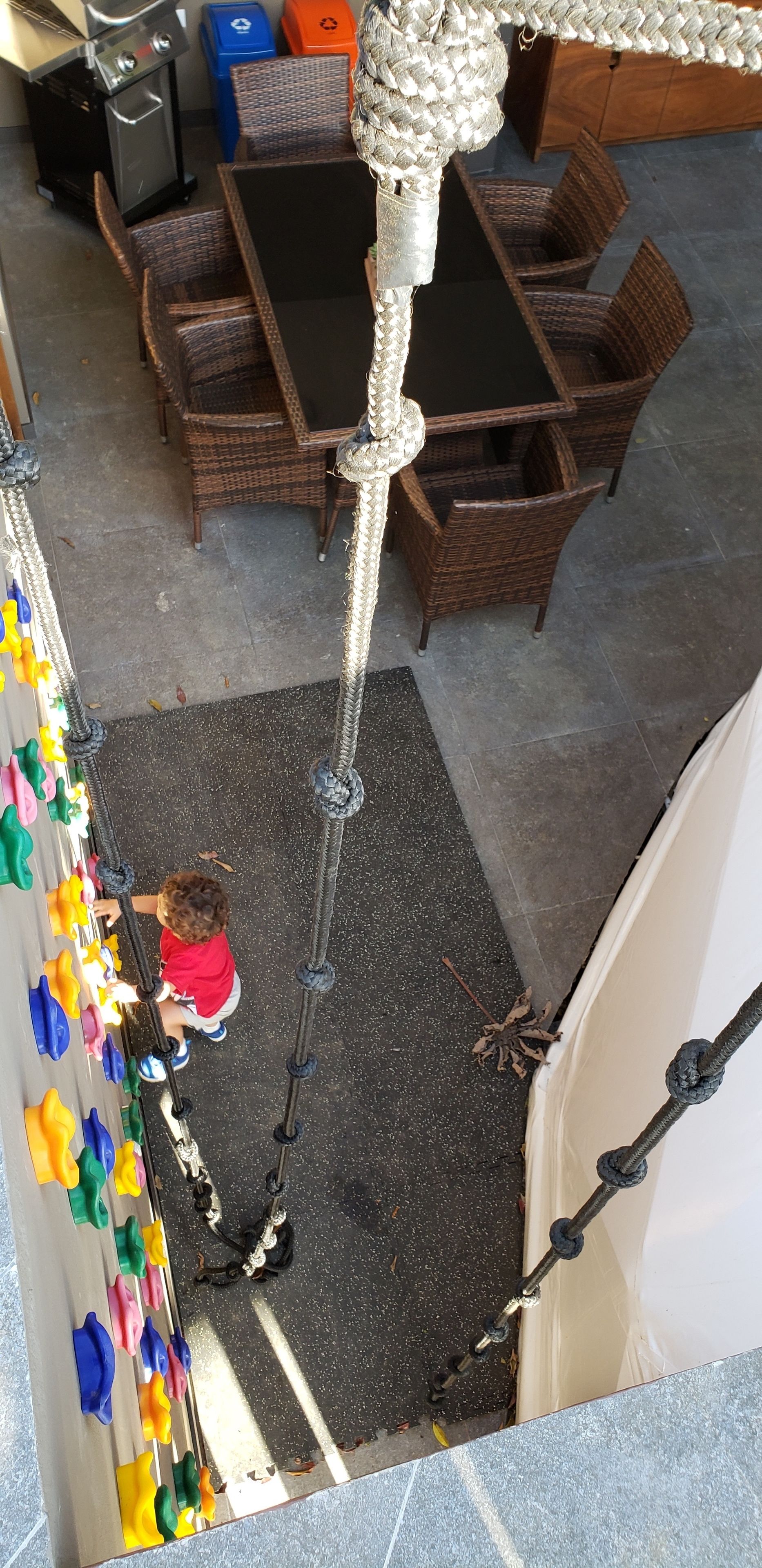 Een kind klimt op een touwladdertje in een kamer met rieten meubels en een zwarte tafel.