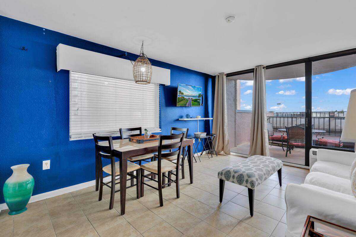 A dining area with a table and chairs, a TV mounted on the wall, and a balcony with outdoor seating.