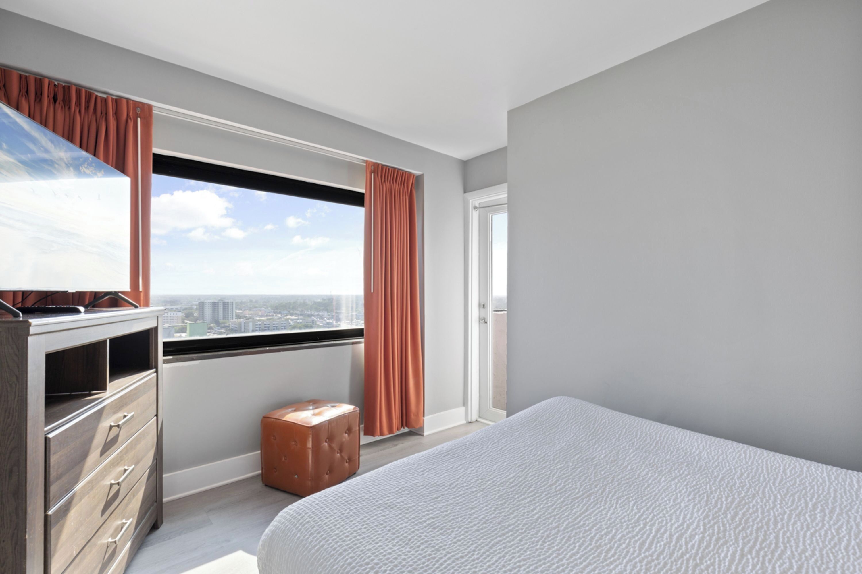 A modern hotel room with a large bed, a dresser, a small ottoman, and a window offering a city view.