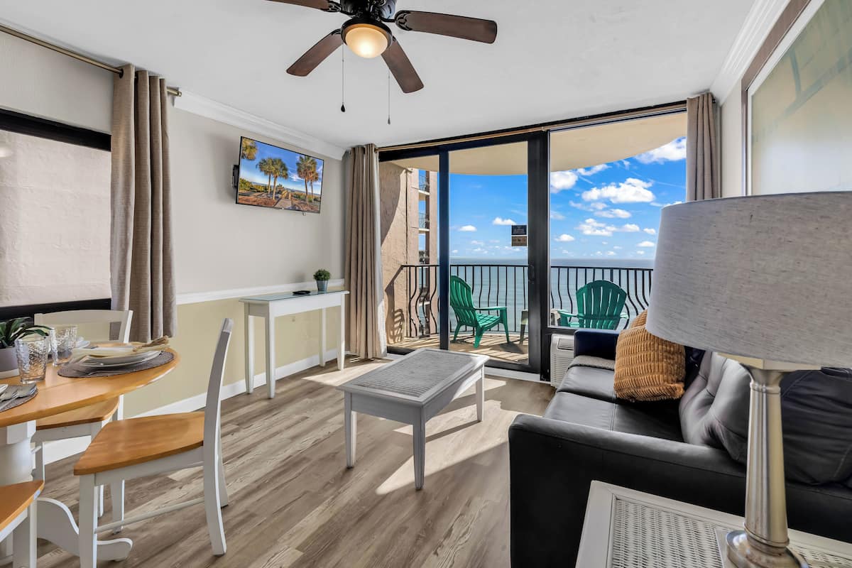 A room with a dining table, chairs, a sofa, a ceiling fan, a TV, and a balcony with a view of the sea.