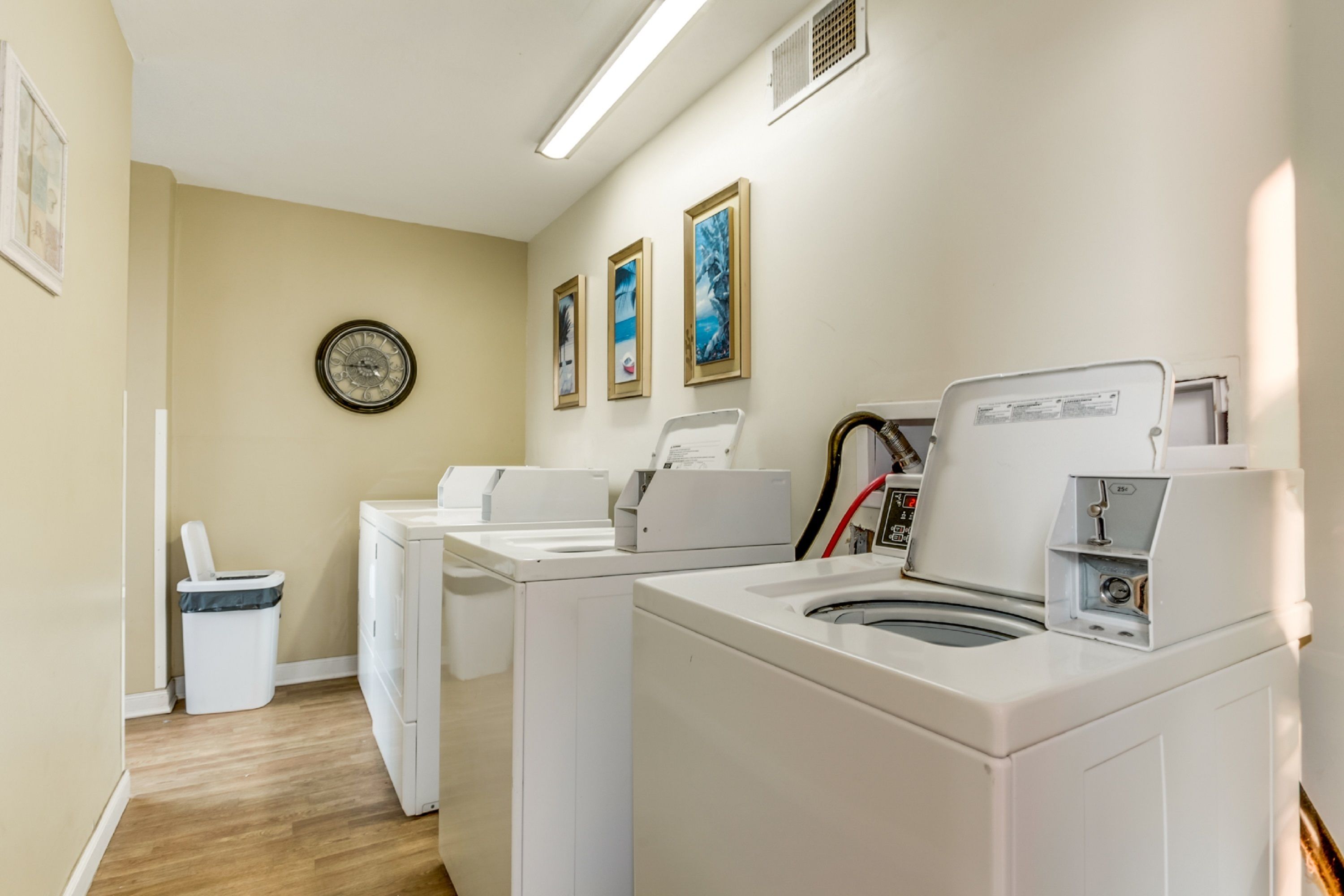 Une buanderie équipée de deux lave-linge et deux sèche-linge à chargement frontal, d’une poubelle et de trois tableaux encadrés accrochés au mur.