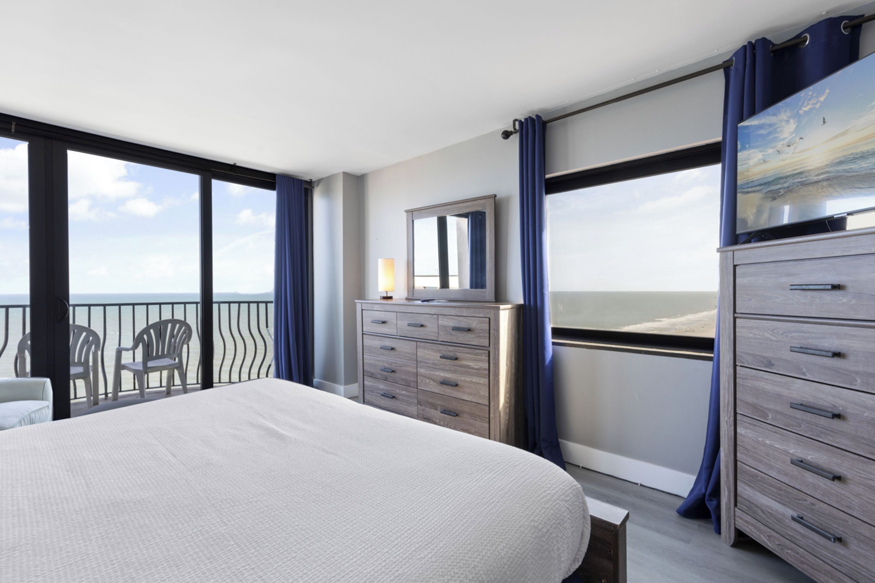 A hotel room with a large bed, a dresser, a mirror, and a view of the ocean through large windows.