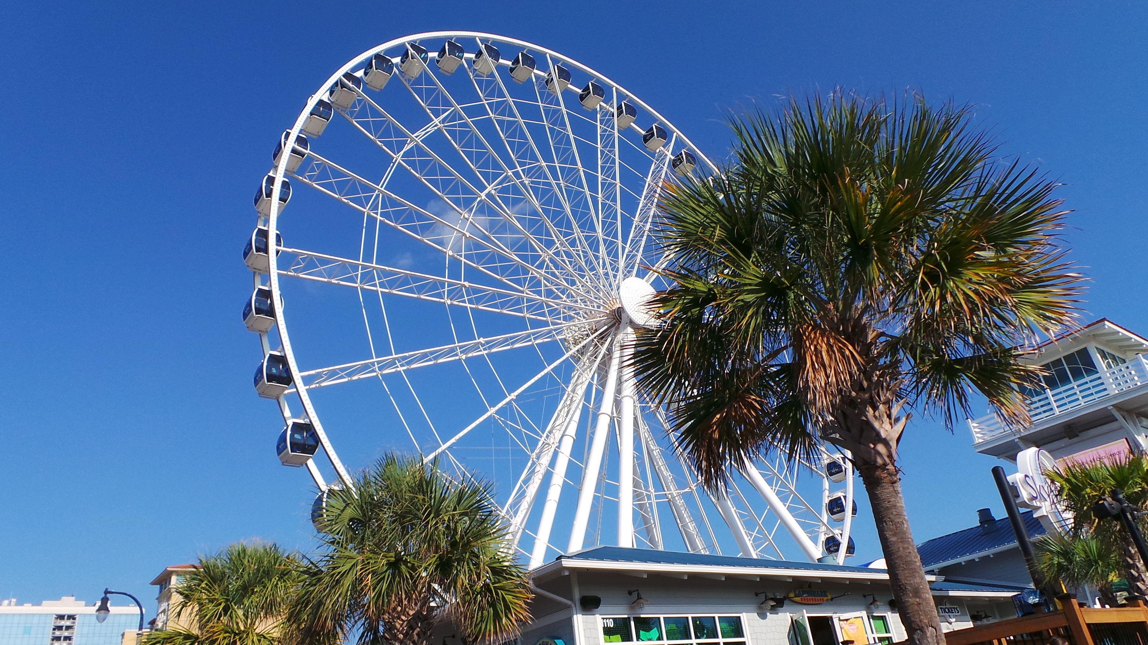 A large Ferris wheel with cabins, palm trees, and a building with a sign.