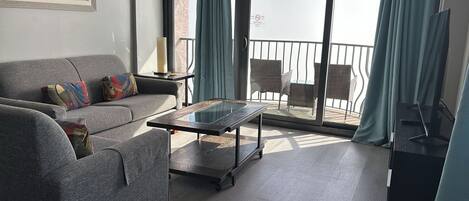 Una sala de estar moderna con un sofá gris, cojines coloridos, una mesa de centro de vidrio y vistas al balcón con muebles de mimbre.