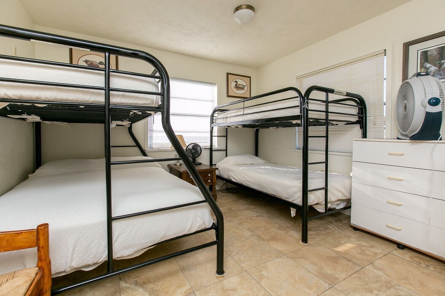 Une chambre de dortoir avec des lits superposés, une commode, un ventilateur et un tableau encadré au mur.