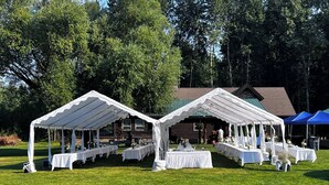 A tented outdoor event space with tables set up for dining, surrounded by a grassy area and trees.