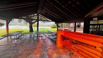 An outdoor covered patio with picnic tables, a barbecue grill, and a wooden bench.