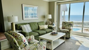 Ein Wohnzimmer mit einem grünen Sofa, einem blumigen Sessel und einem weißen Couchtisch. Ein großes Fenster bietet Blick auf Strand und Meer.