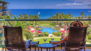 A balcony with wicker chairs and a table, overlooking a pool and a view of the ocean.