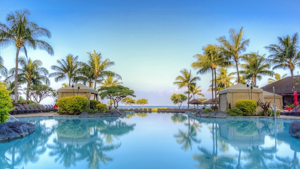 A tranquil pool area with clear blue water, surrounded by palm trees and lounge chairs, reflecting the sky and nearby structures.