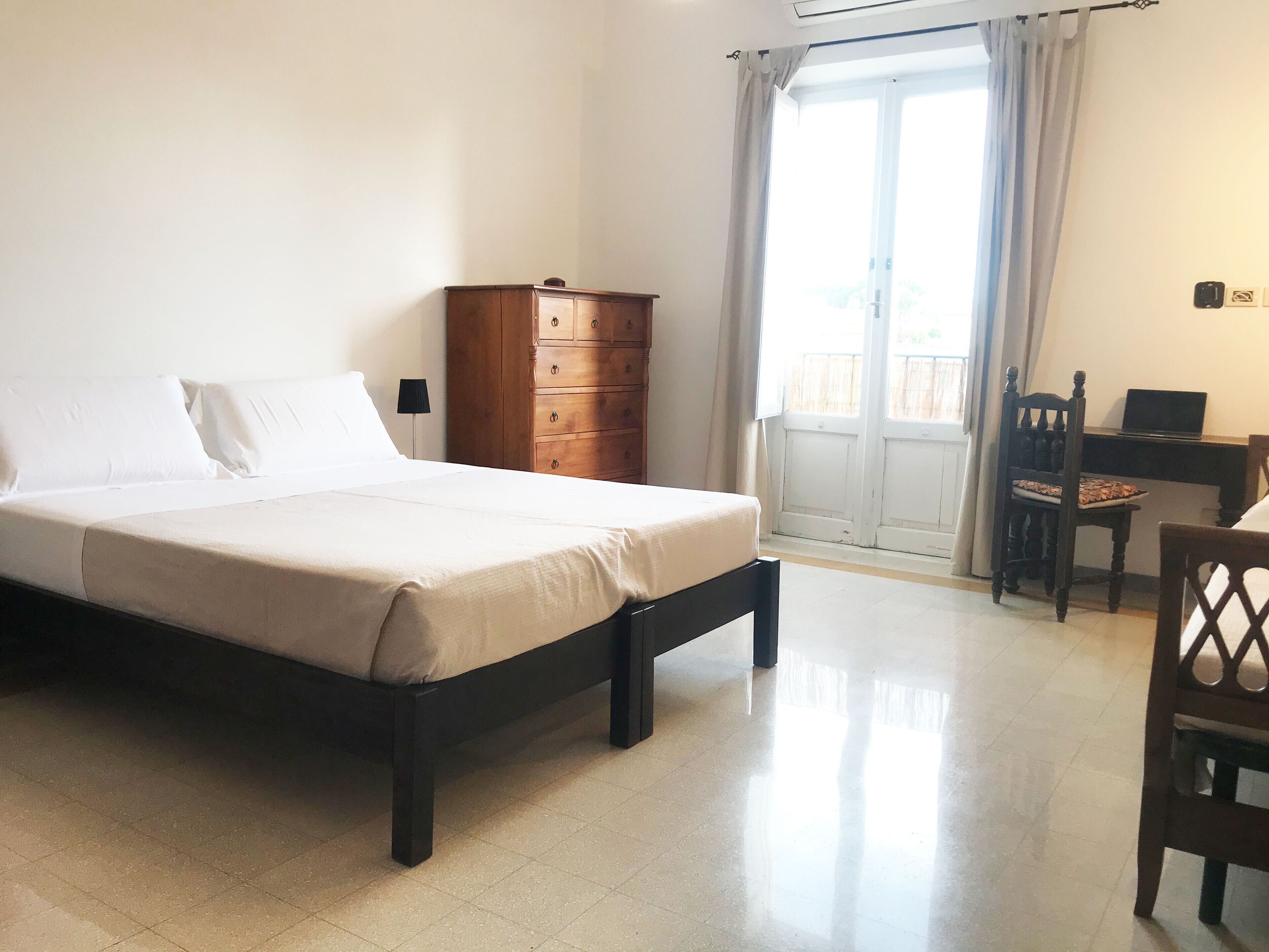 A neatly made bed with a wooden headboard, a wooden dresser, a desk with a chair, and a window with curtains.