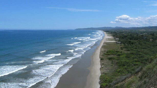 Eine Küstenlandschaft mit Sandstrand, Wellen und fernen Hügeln.