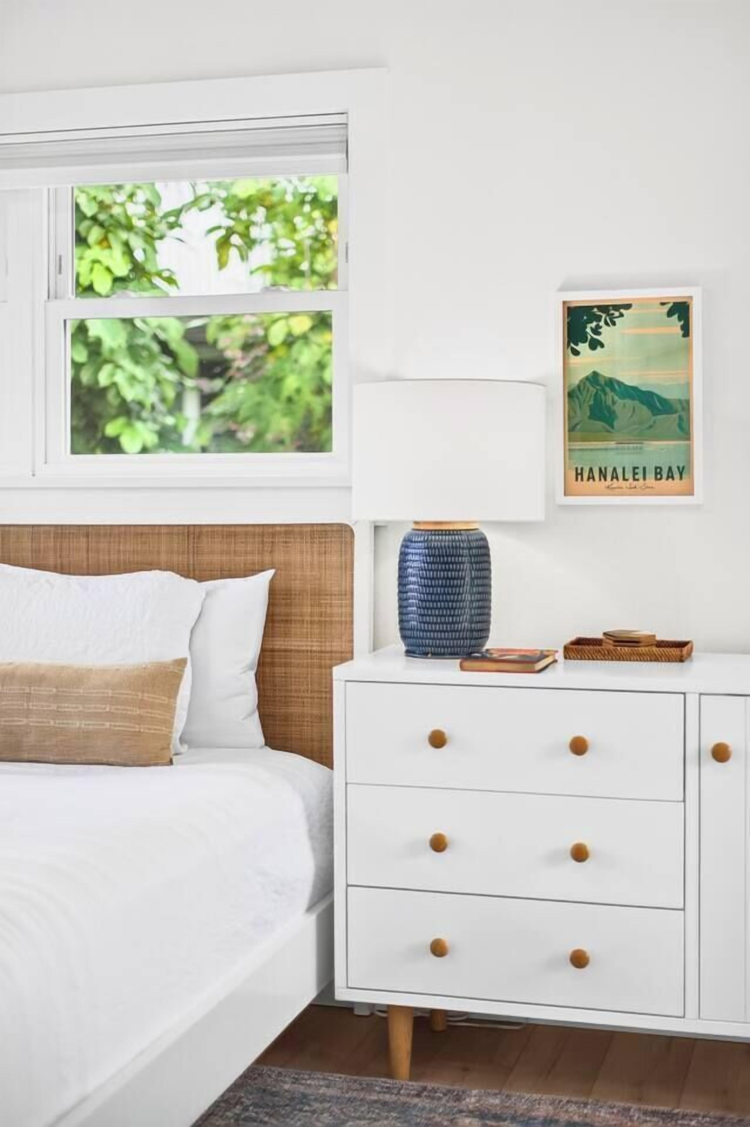 Une chambre à coucher avec un lit, une table de chevet, un tableau encadré et une fenêtre donnant sur un paysage verdoyant.