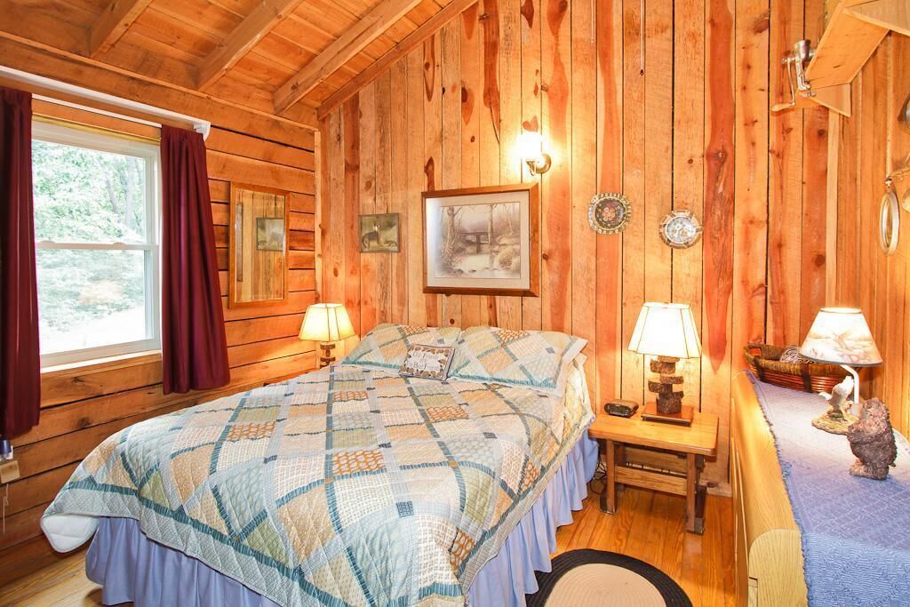Un dormitorio de cabaña de madera con una cama, dos lámparas de noche, un cuadro en la pared y una ventana con cortinas.