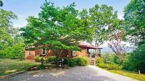 A wooden cabin with a gravel driveway, surrounded by lush greenery and trees.