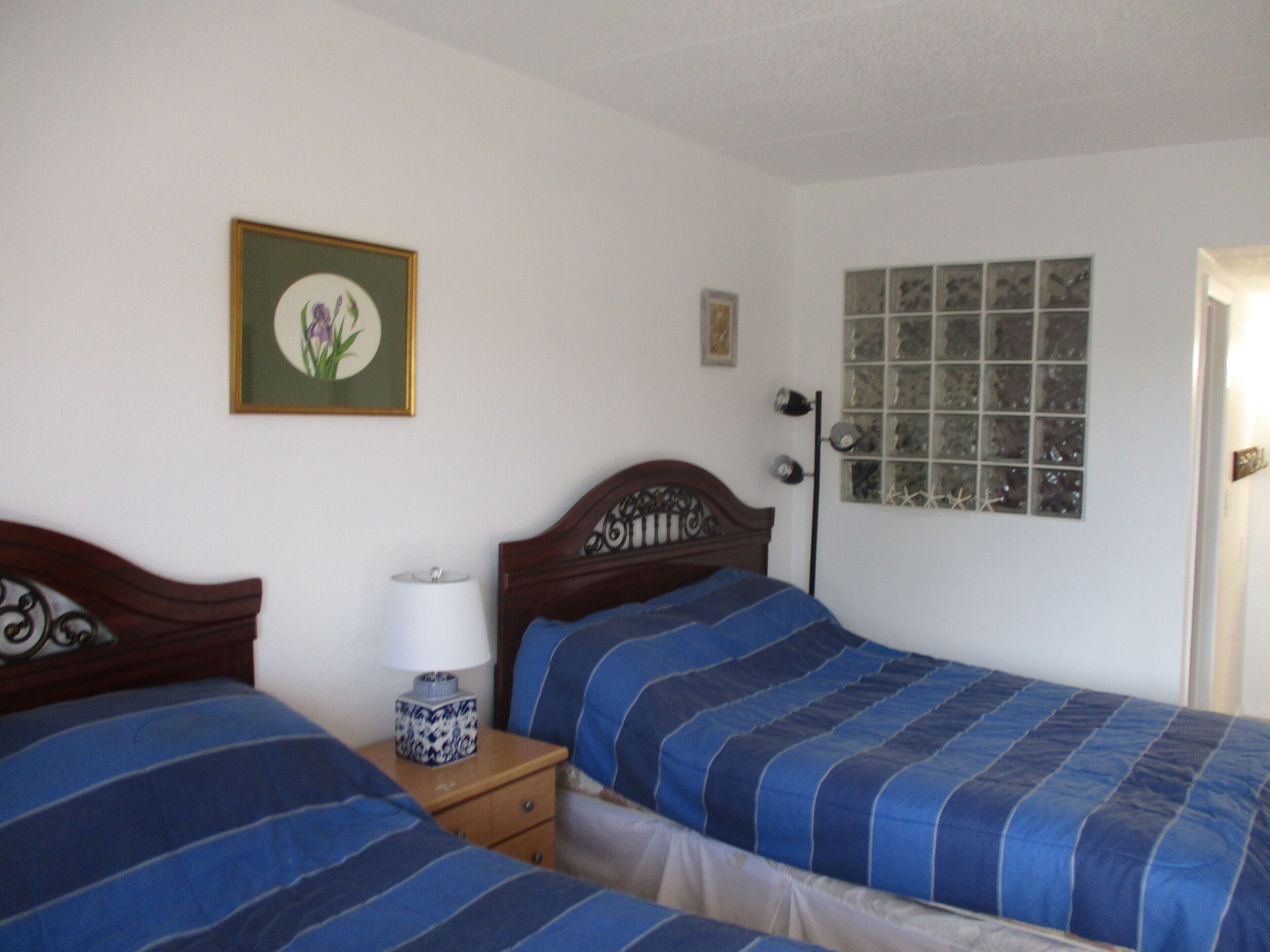 A bedroom with two single beds, a bedside table with a lamp, a framed picture on the wall, and a window with a view of trees.