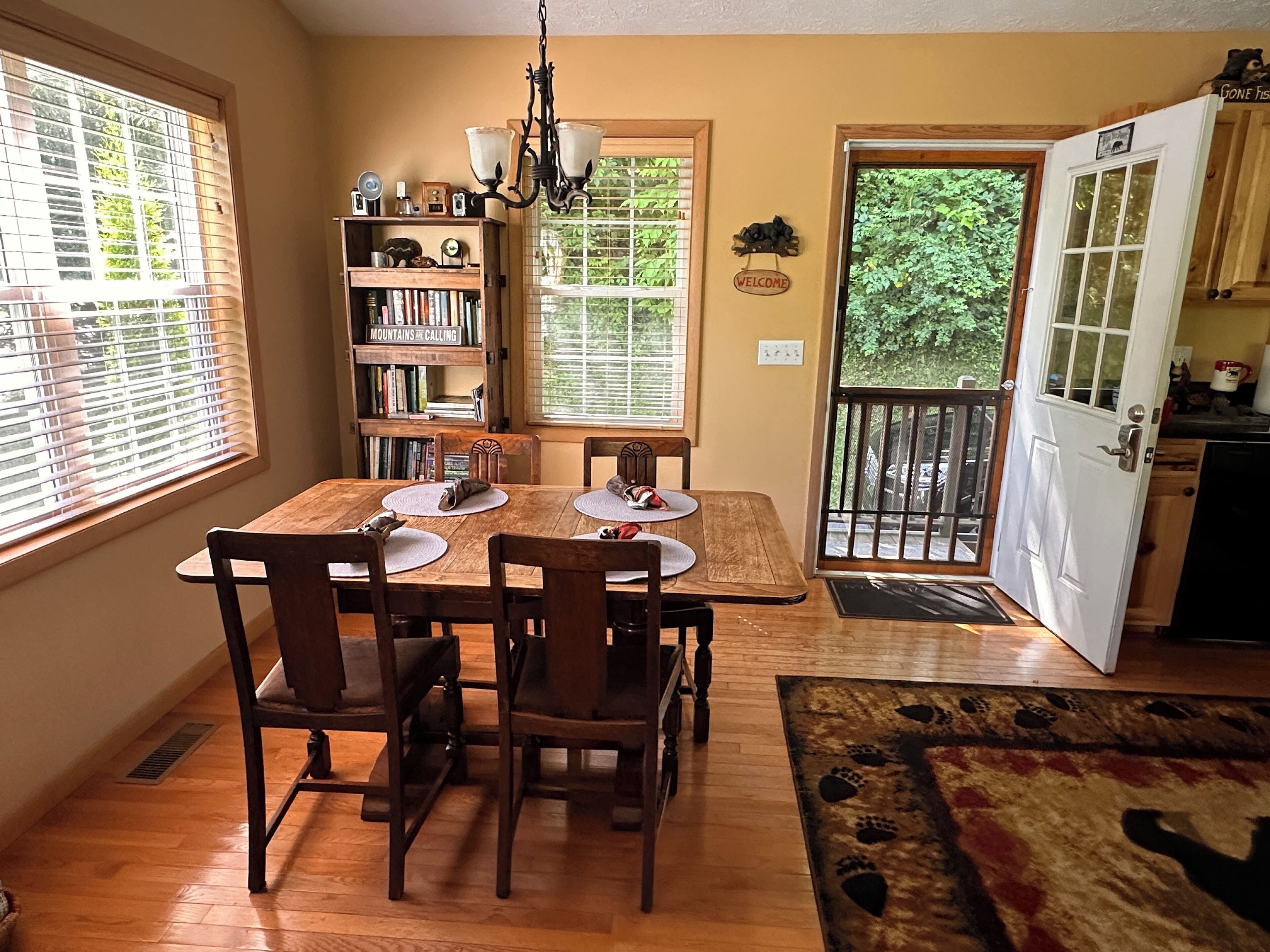 A dining area with a wooden table, four chairs, a chandelier, and a bookshelf.