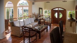 Un vaste espace de vie comprenant un canapé, des fauteuils et une table basse. De grandes fenêtres et une porte au motif circulaire s’y trouvent.