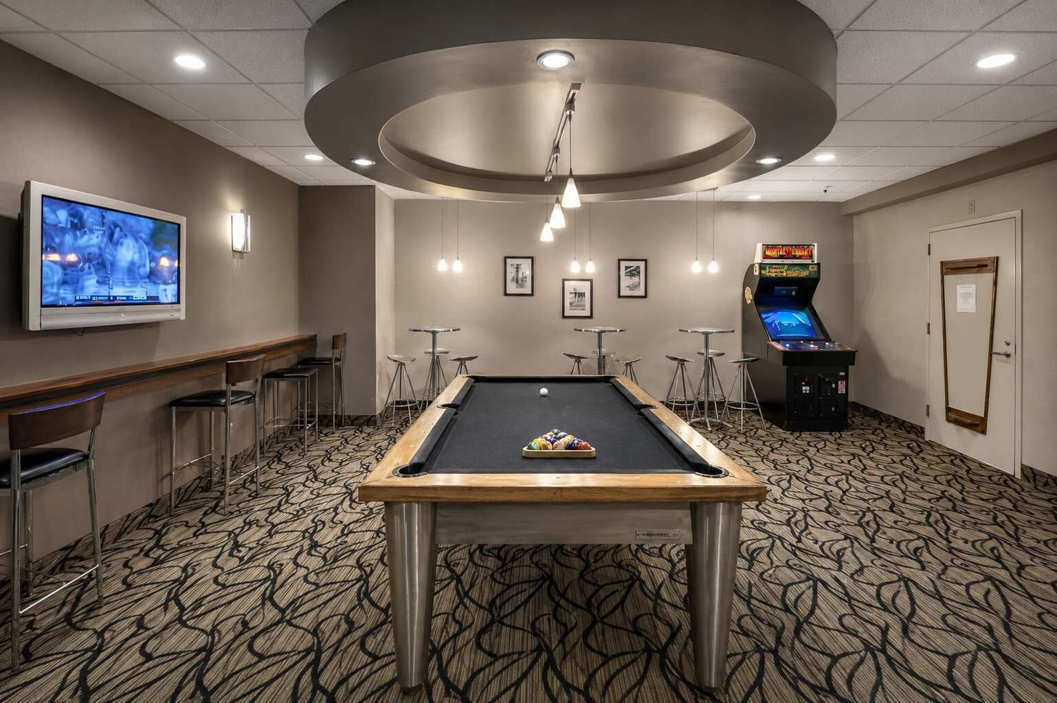 Une table de billard avec un queue de billard, un téléviseur fixé au mur et une machine à pinces dans une pièce dont le sol est recouvert d’une moquette à motifs et qui est équipée de tabourets de bar.
