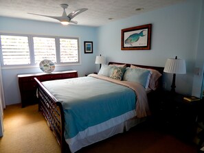 A bedroom with a bed, a ceiling fan, a window with blinds, a framed picture, a nightstand with a lamp, and a small table with a decorative plate. - Must see! Spectacular Direct Oceanfront Corner Unit  (Kailua-Kona)