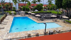 A swimming pool with stone tiles, surrounded by lounge chairs and umbrellas, with a view of a red-roofed building and palm trees in the background. - Must see! Spectacular Direct Oceanfront Corner Unit  (Kailua-Kona)