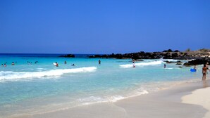 A beach with clear turquoise water, people swimming and surfing, and rocky outcrops in the distance. - Must see! Spectacular Direct Oceanfront Corner Unit  (Kailua-Kona)