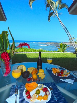 Una mesa con frutas tropicales, una botella de champán y un vaso de jugo de naranja, con vista a una playa con palmeras y el océano.