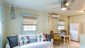 A cozy living area with a sofa, a dining table, wicker chairs, a ceiling fan, and a refrigerator.