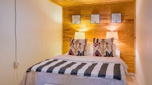 A bedroom with a wooden ceiling, a bed with striped bedding, two wall-mounted lamps, and three framed pictures on the wall.