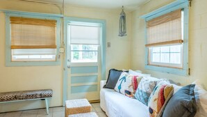 A room with a white sofa, a bench, and two wicker poufs. There are two windows with blinds, one on each side of the door.