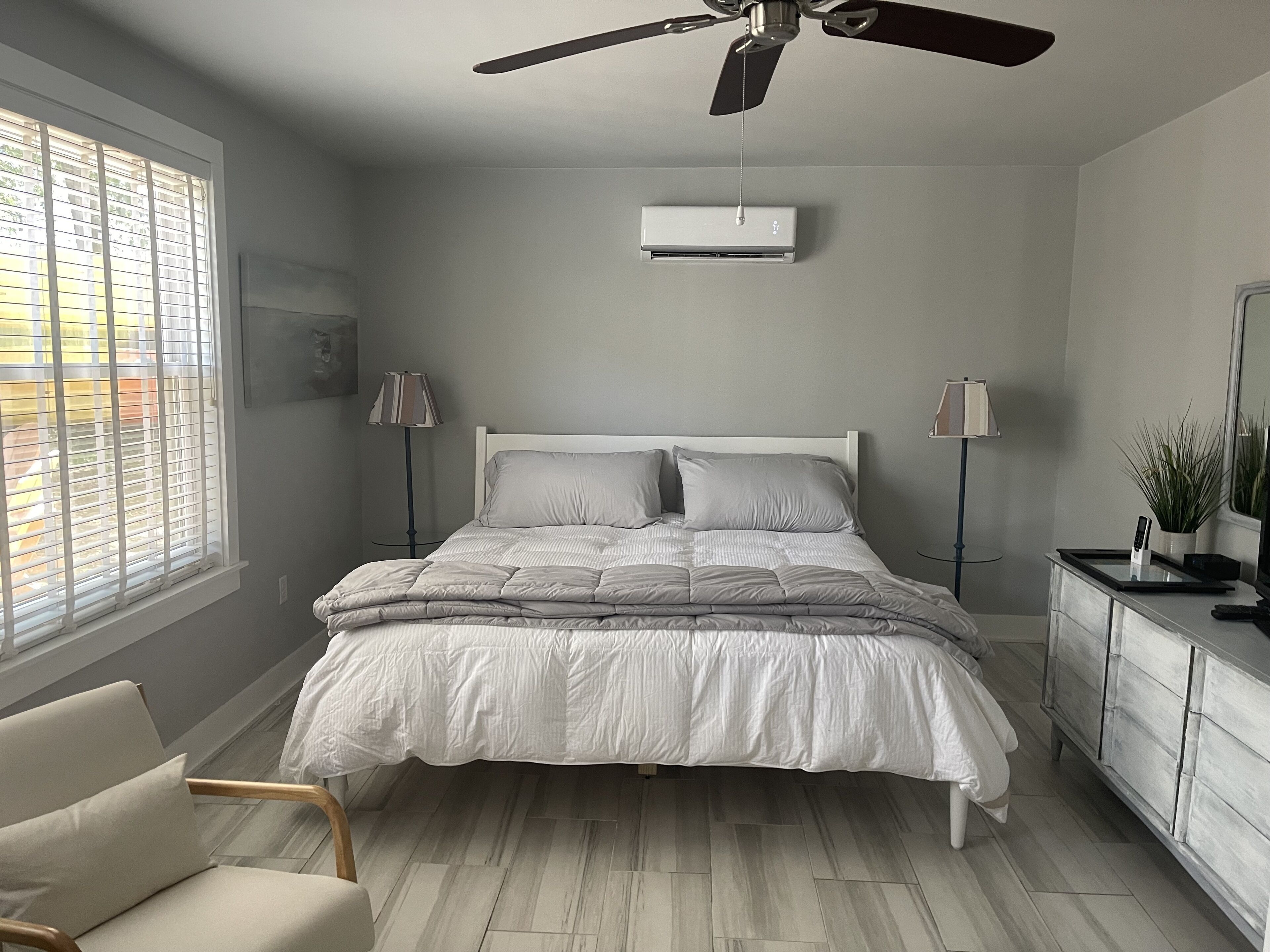 Un dormitorio con cama, ventilador de techo, aire acondicionado, cómoda, silla y ventana con persianas.