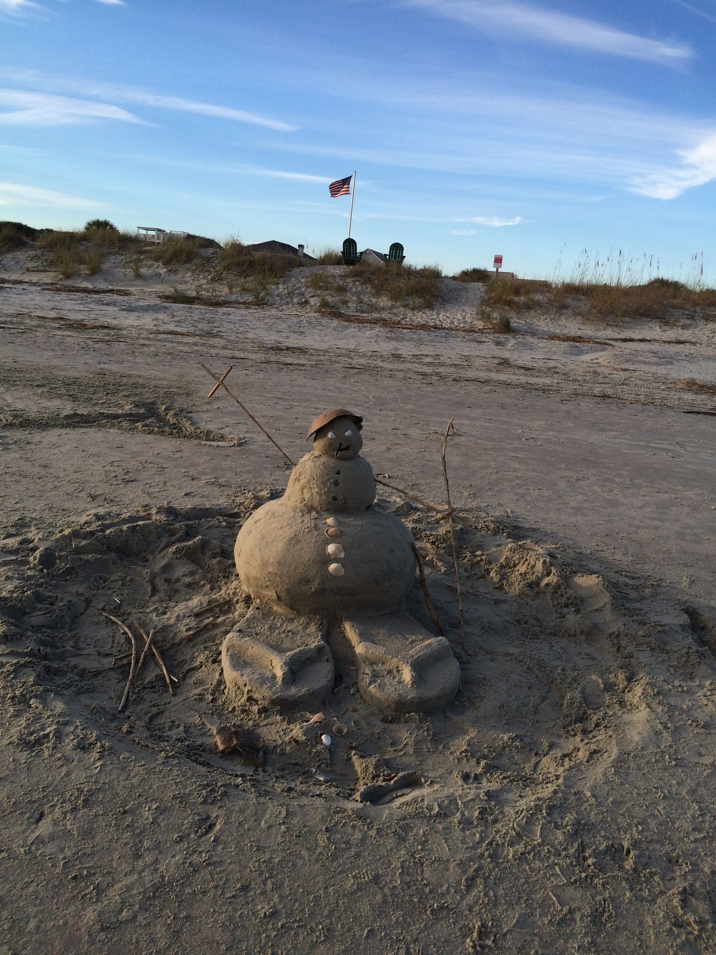 A sand sculpture of a snowman with a flag on top, located on a sandy beach with sparse vegetation and a clear sky.