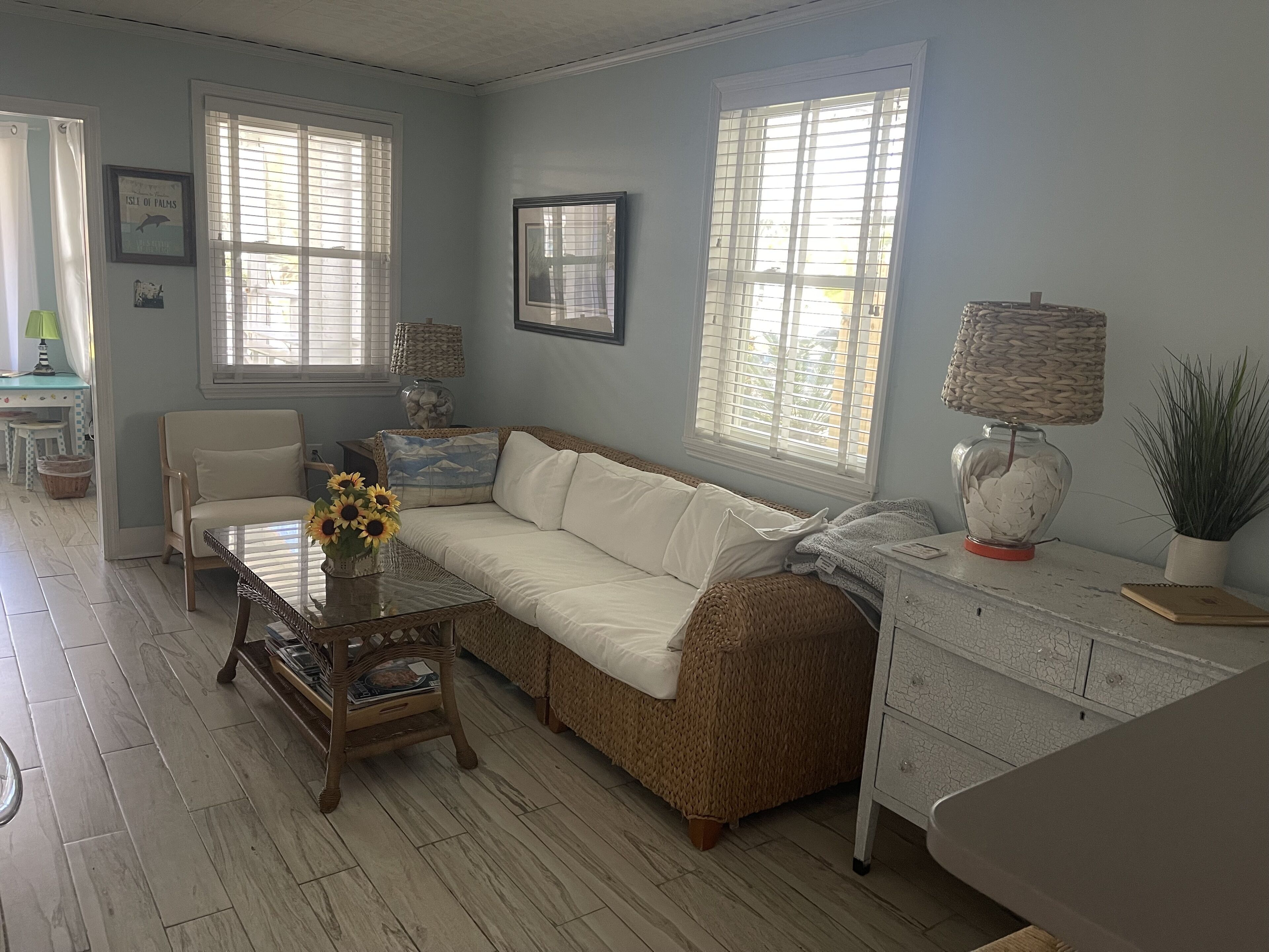 A living room with a sofa, a coffee table with a vase of sunflowers, a wicker chair, and a side table with a lamp and a plant.
