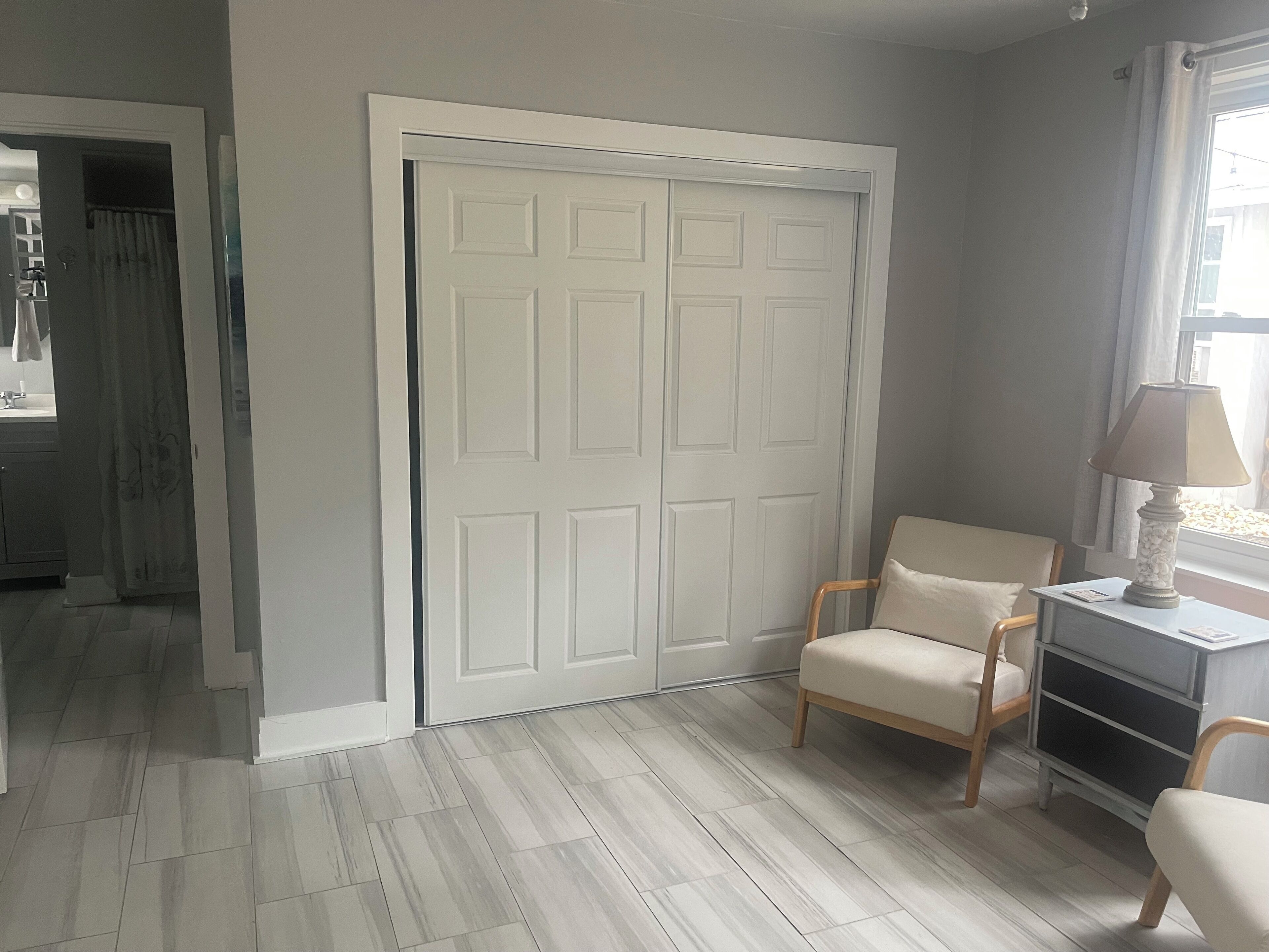 Una habitación con una puerta blanca, una silla, una mesa con lámpara y una ventana con cortinas.