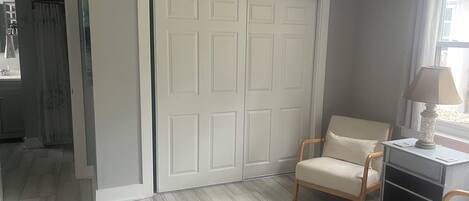 Una habitación con una puerta blanca, una silla, una mesa con lámpara y una ventana con cortinas.
