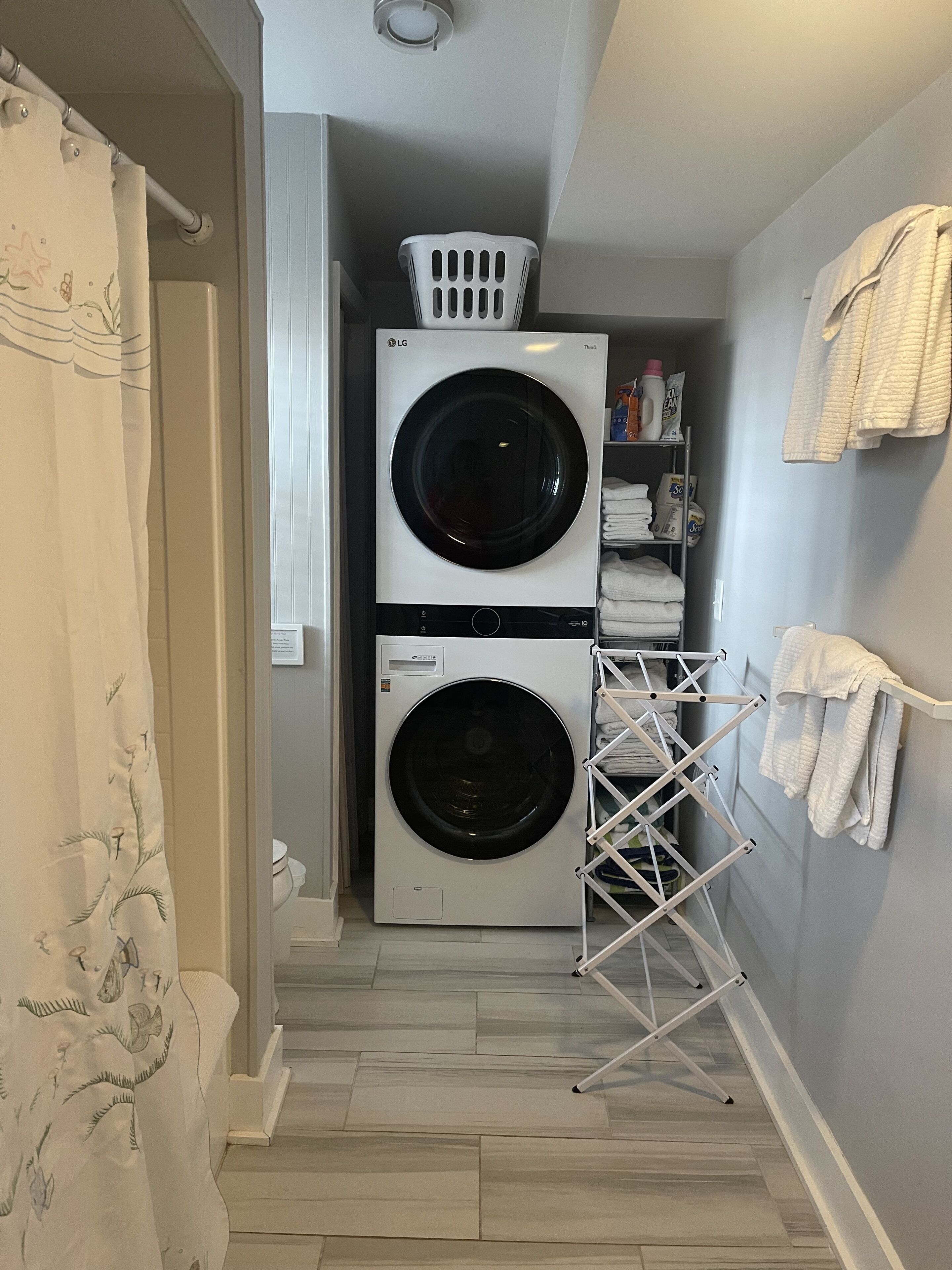 A laundry room with two stacked front-loading washing machines, a drying rack, and a shelf with laundry supplies.