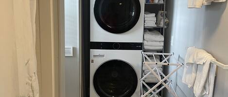 A laundry room with two stacked front-loading washing machines, a drying rack, and a shelf with laundry supplies.