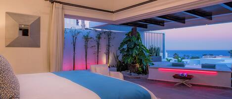A modern bedroom with a large bed, a view of the ocean, and a balcony with plants and a red light.