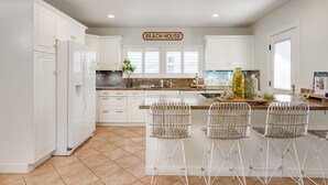 Uma cozinha com armários brancos, uma ilha central com banquetas, uma placa com a inscrição "BEACH HOUSE" e uma janela grande com persianas.