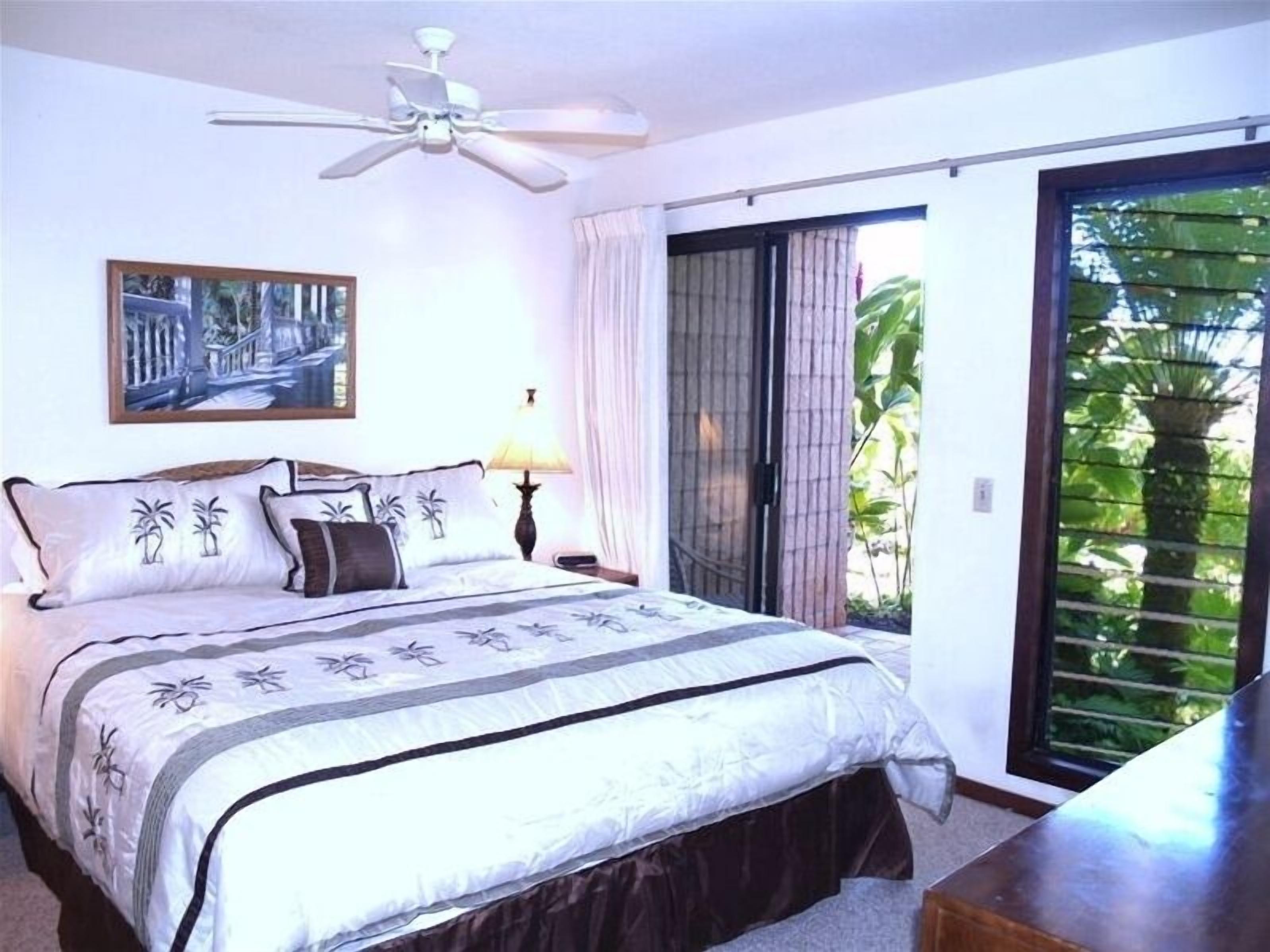Une chambre à coucher comprenant un lit, un ventilateur de plafond, une table de chevet avec une lampe, un tableau au mur et une porte-fenêtre coulissante donnant sur un balcon agrémenté de plantes.