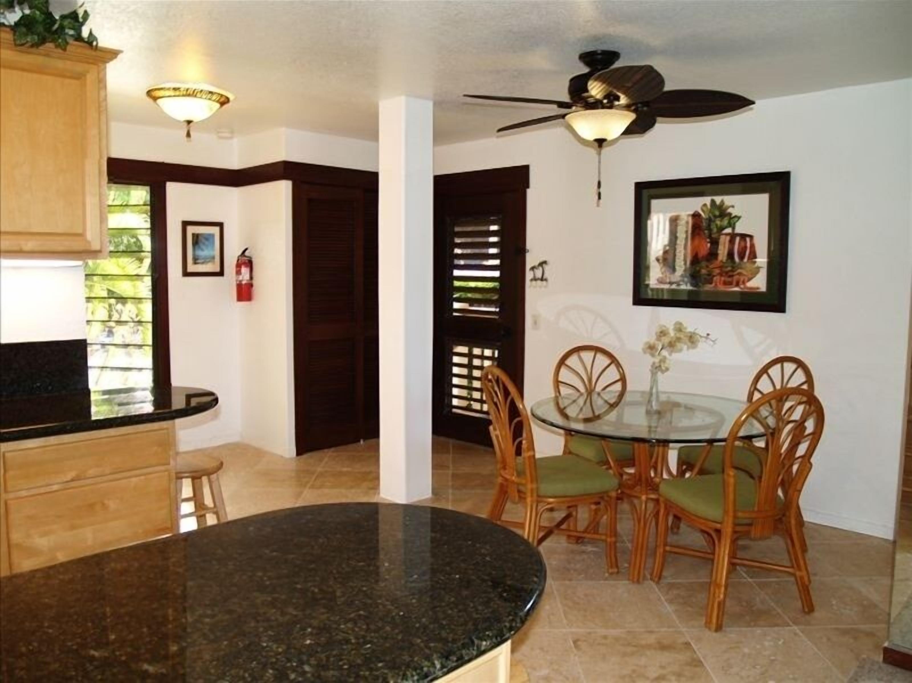A kitchen with a granite countertop, wooden cabinets, a ceiling fan, and a dining area with a glass table and wicker chairs.