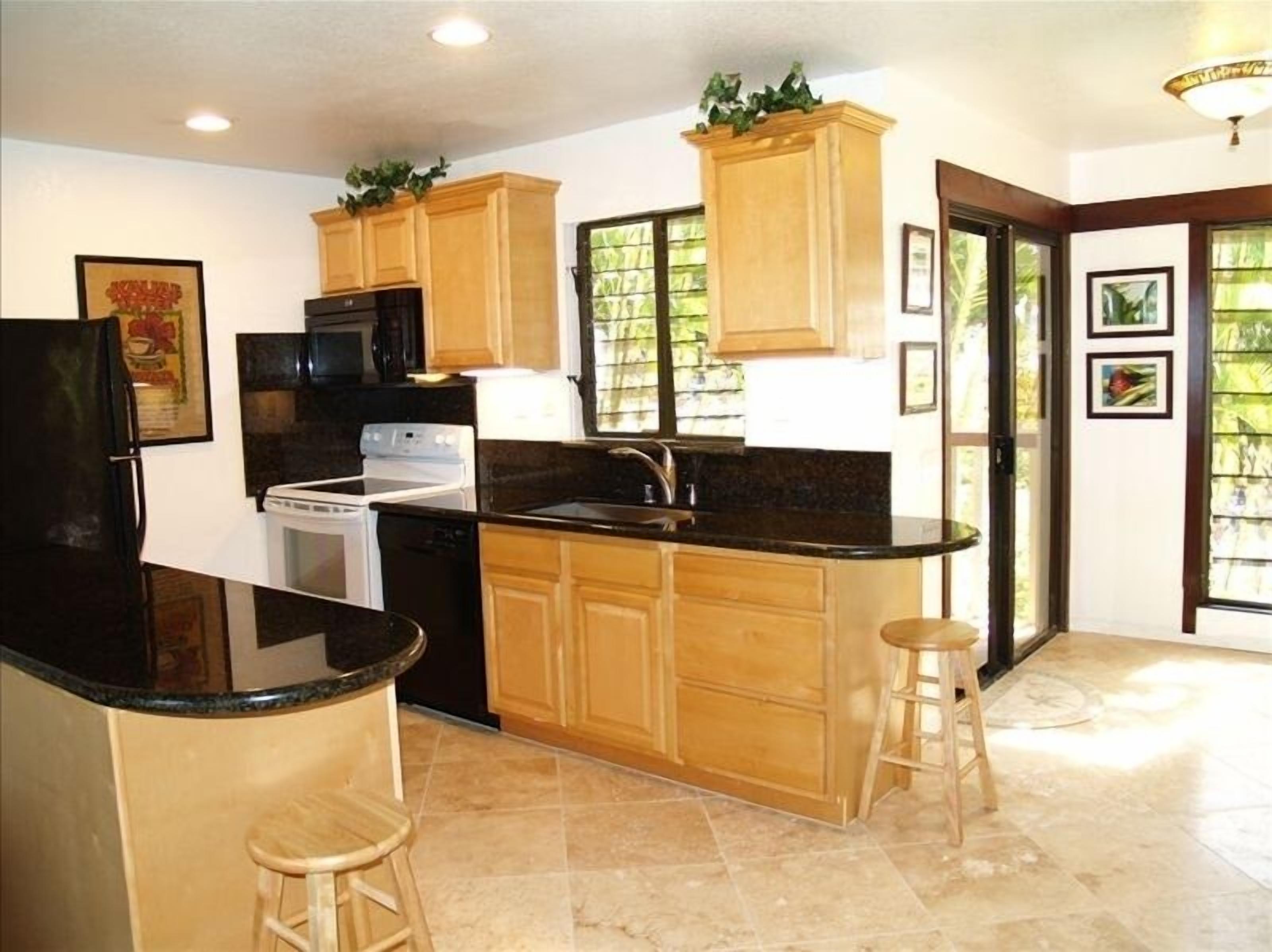 A kitchen with wooden cabinets, a black countertop, a stove, and a refrigerator. There are two stools and a window with a view of greenery.