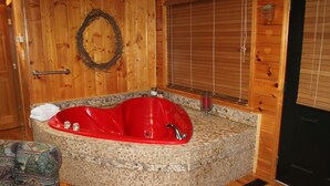 A wooden cabin interior with a red jacuzzi tub, a woven wreath on the wall, and a window with blinds. - Cozy & Perfect For A Romantic Getaway For Two! (Pigeon Forge)