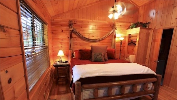 A wooden cabin bedroom with a bed, a nightstand, a lamp, and a hammock. - Cozy & Perfect For A Romantic Getaway For Two! (Pigeon Forge)